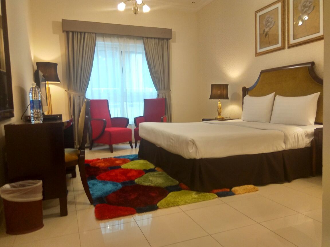 Habitación de hotel con cama, una silla, escritorio, lámpara, ventana con cortinas y una alfombra colorida.