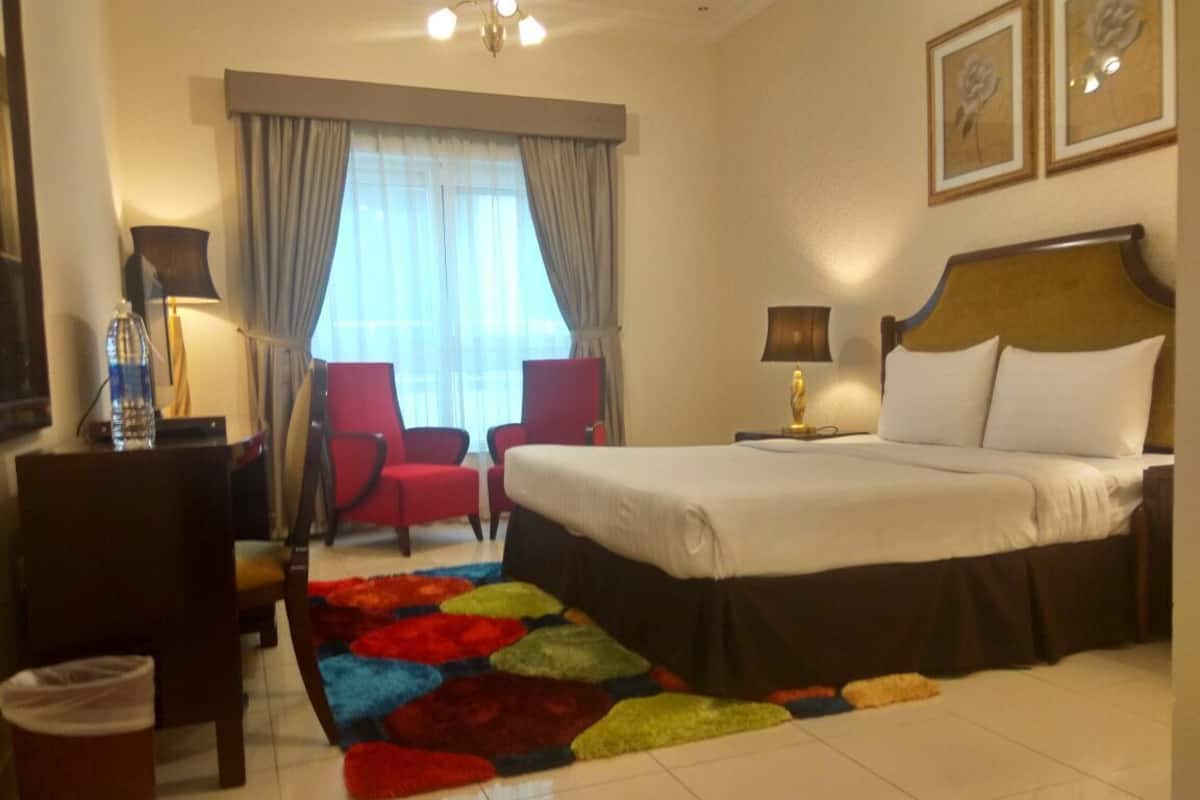 A hotel room with a bed, a chair, a desk, a lamp, a window with curtains, and a colorful rug.