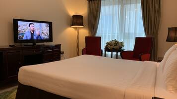 A hotel room with a large bed, a television on a dresser, two chairs, a small table with a flower arrangement, and a window with curtains.