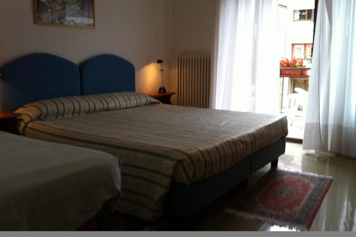 A bedroom with a large bed, a smaller bed, a nightstand with a lamp, a radiator, and a window with curtains.