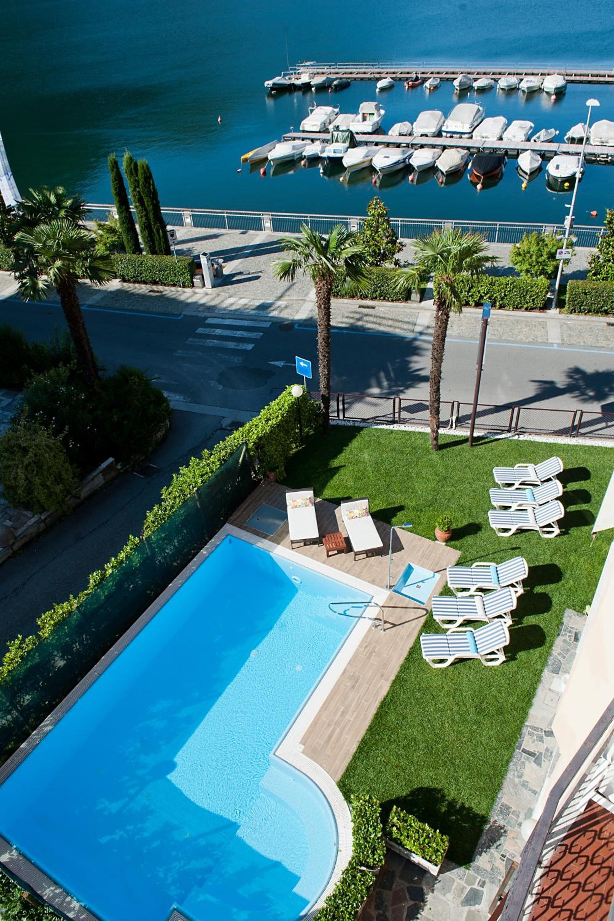 Une piscine avec des chaises longues, un port de plaisance avec des bateaux et une route bordée de palmiers.