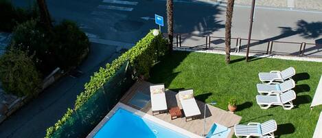 Une piscine avec des chaises longues, un port de plaisance avec des bateaux et une route bordée de palmiers.