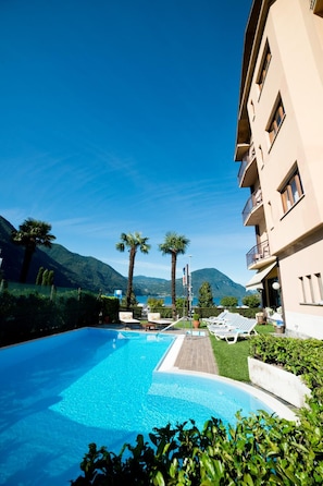 Una piscina con tumbonas, un edificio con balcones y un paisaje montañoso al fondo.