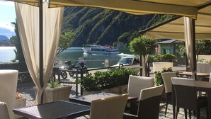 Un espace extérieur aménagé avec des tables et des chaises, offrant une vue sur un plan d’eau où naviguent des bateaux, le tout sur fond de montagnes.