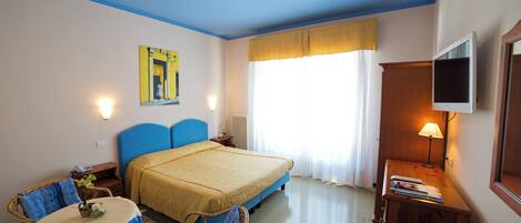 A hotel room with a bed, a desk, a chair, a table with a plant, a lamp, and a television.