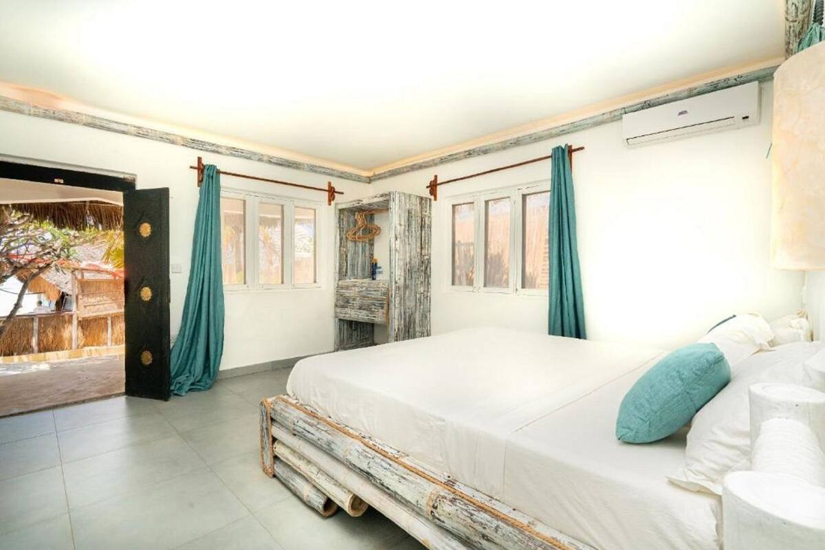 Une chambre dotée d’un grand lit, d’une tête de lit en bois rustique, de rideaux turquoise et offrant une vue sur un espace extérieur tropical.