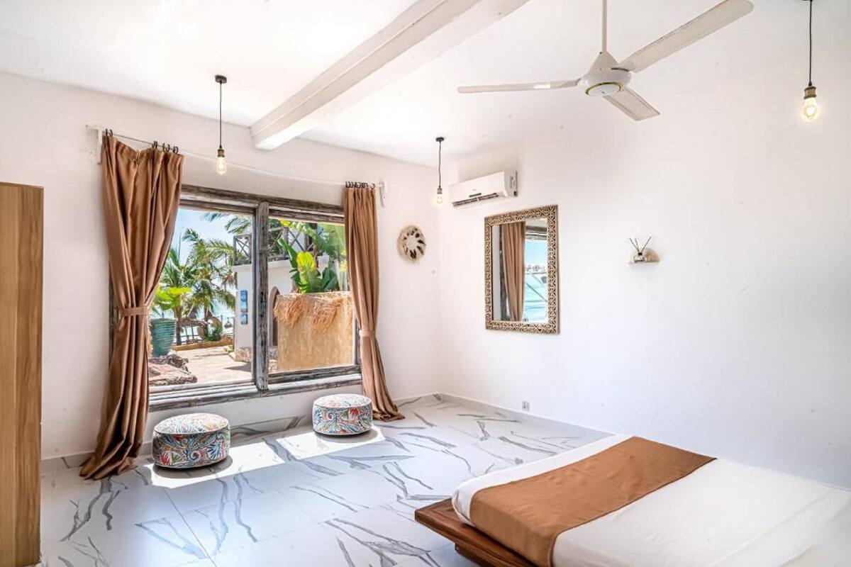 Une chambre à coucher comprenant un lit, un ventilateur de plafond, une fenêtre avec des rideaux, une petite table ronde et un miroir décoratif.