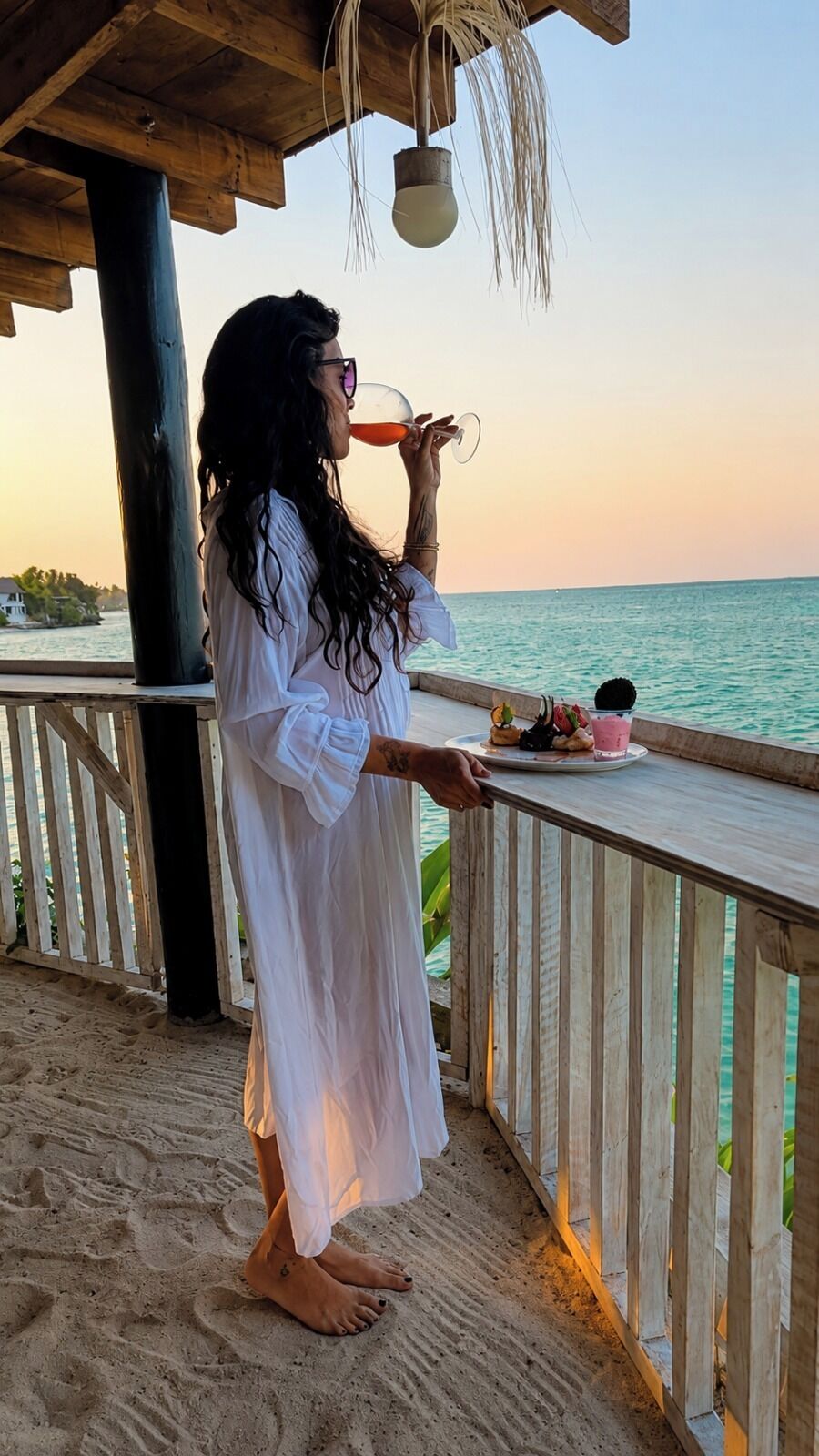 A person standing on a wooden deck overlooking a body of water, with a railing and a table with a drink and food items.
