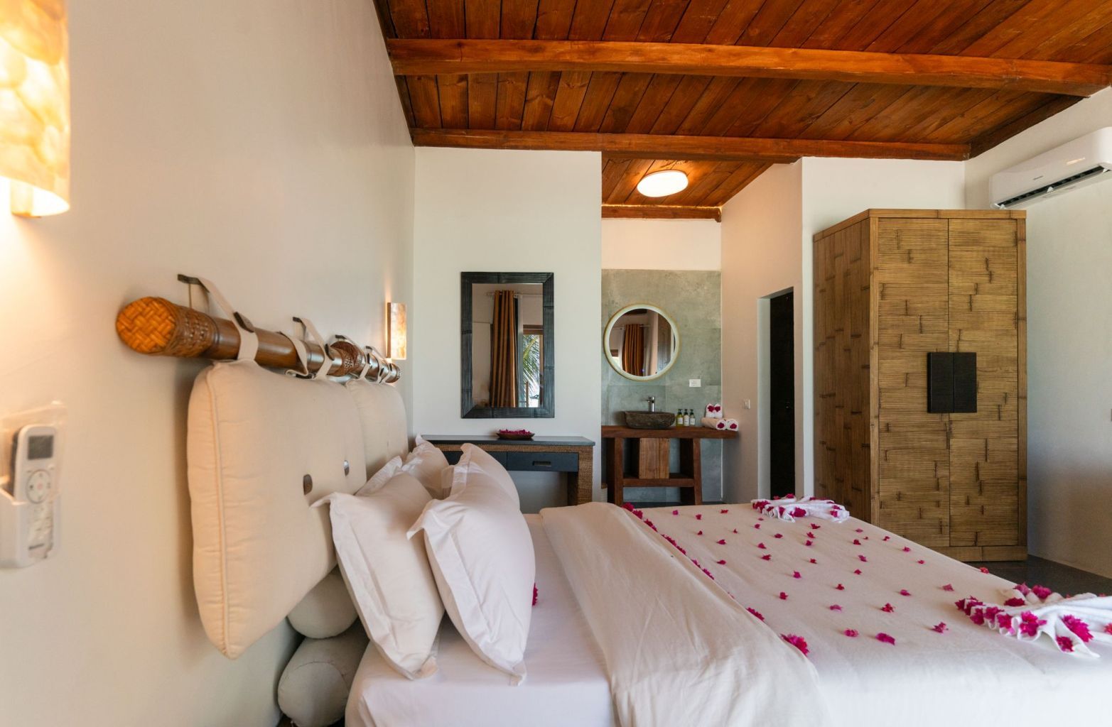 A hotel room with a wooden ceiling, a bed with white linens, a bedside table, a mirror, a wooden wardrobe, and a bathroom with a sink and mirror.