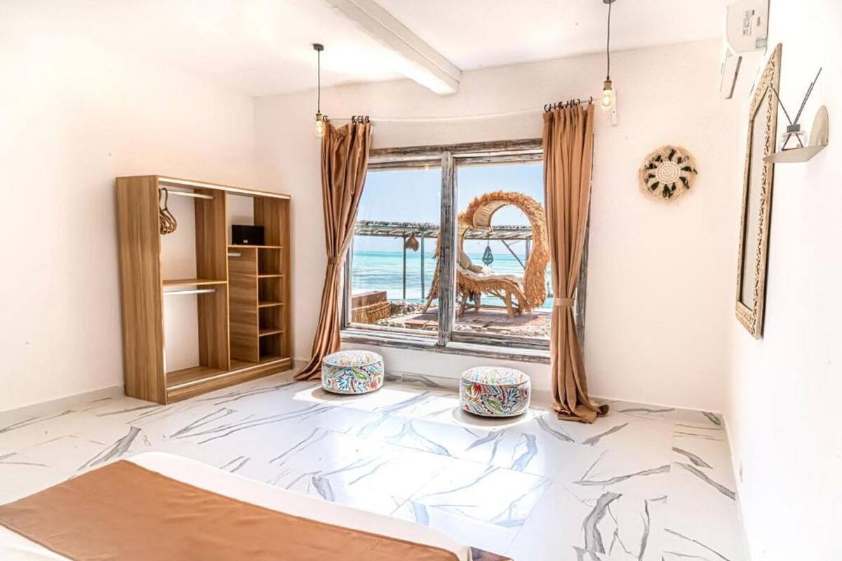 Une chambre avec un lit, une armoire en bois, une fenêtre donnant sur la plage, une chaise en osier et une petite table ronde.