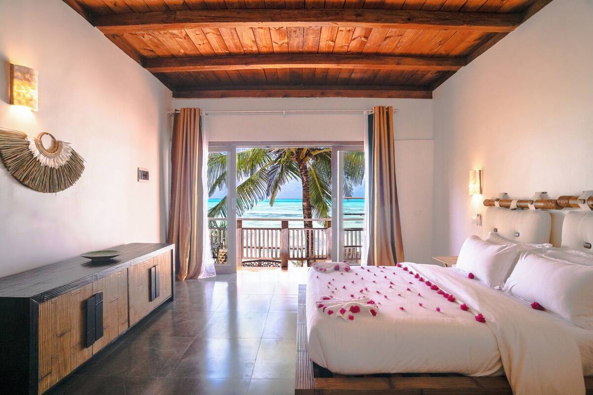 Une chambre d’hôtel avec un plafond en bois, un lit avec une literie blanche et des fleurs roses, une armoire en bois et une vue sur l’océan depuis un balcon.