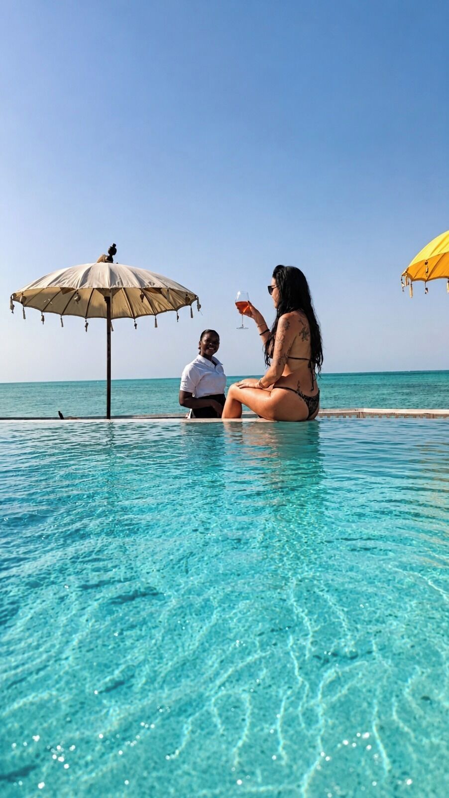 A poolside scene with two people under umbrellas, clear blue water, and a calm sea in the background.