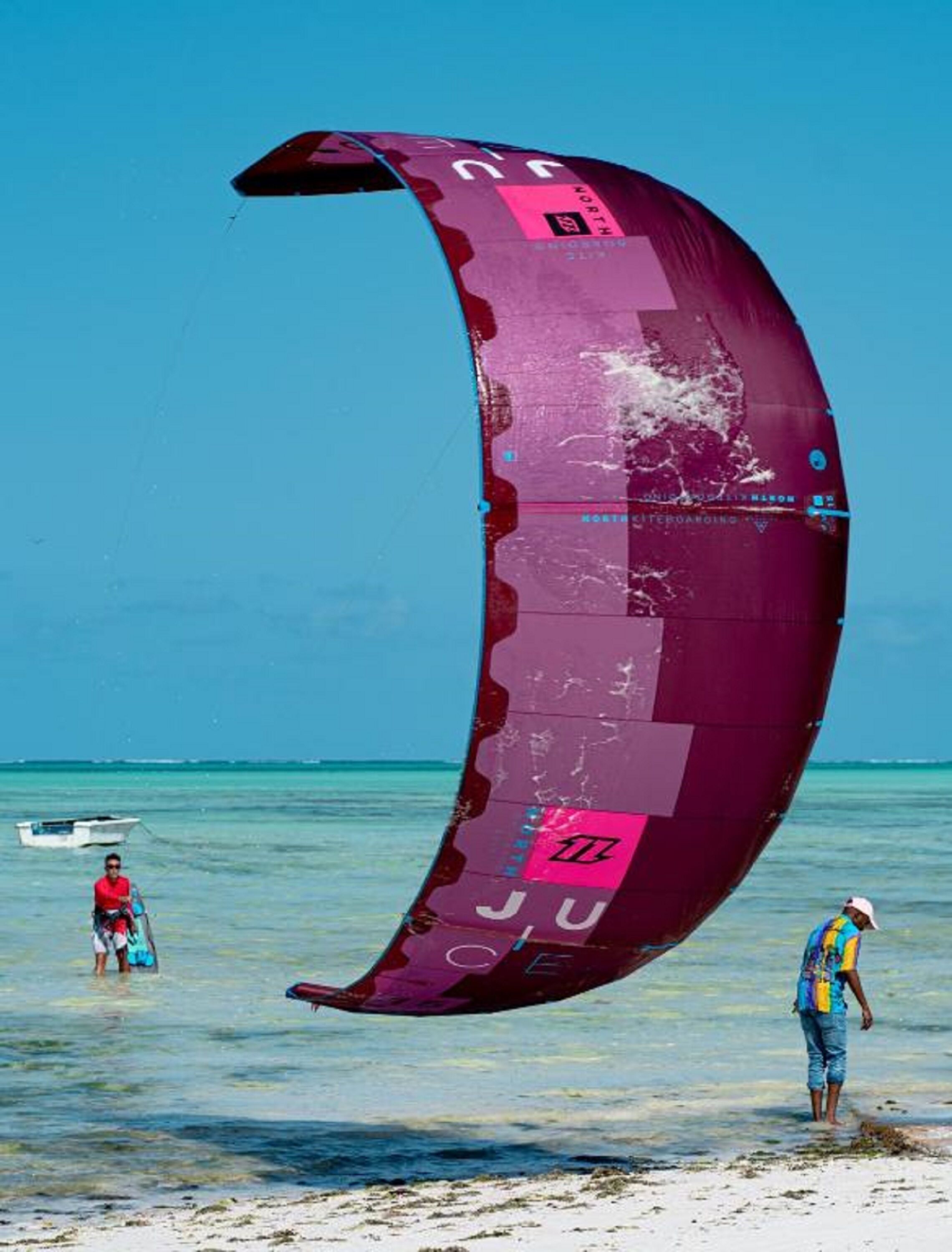 A beach scene with a large purple kiteboard in the water, a small boat, and two people standing in shallow water.