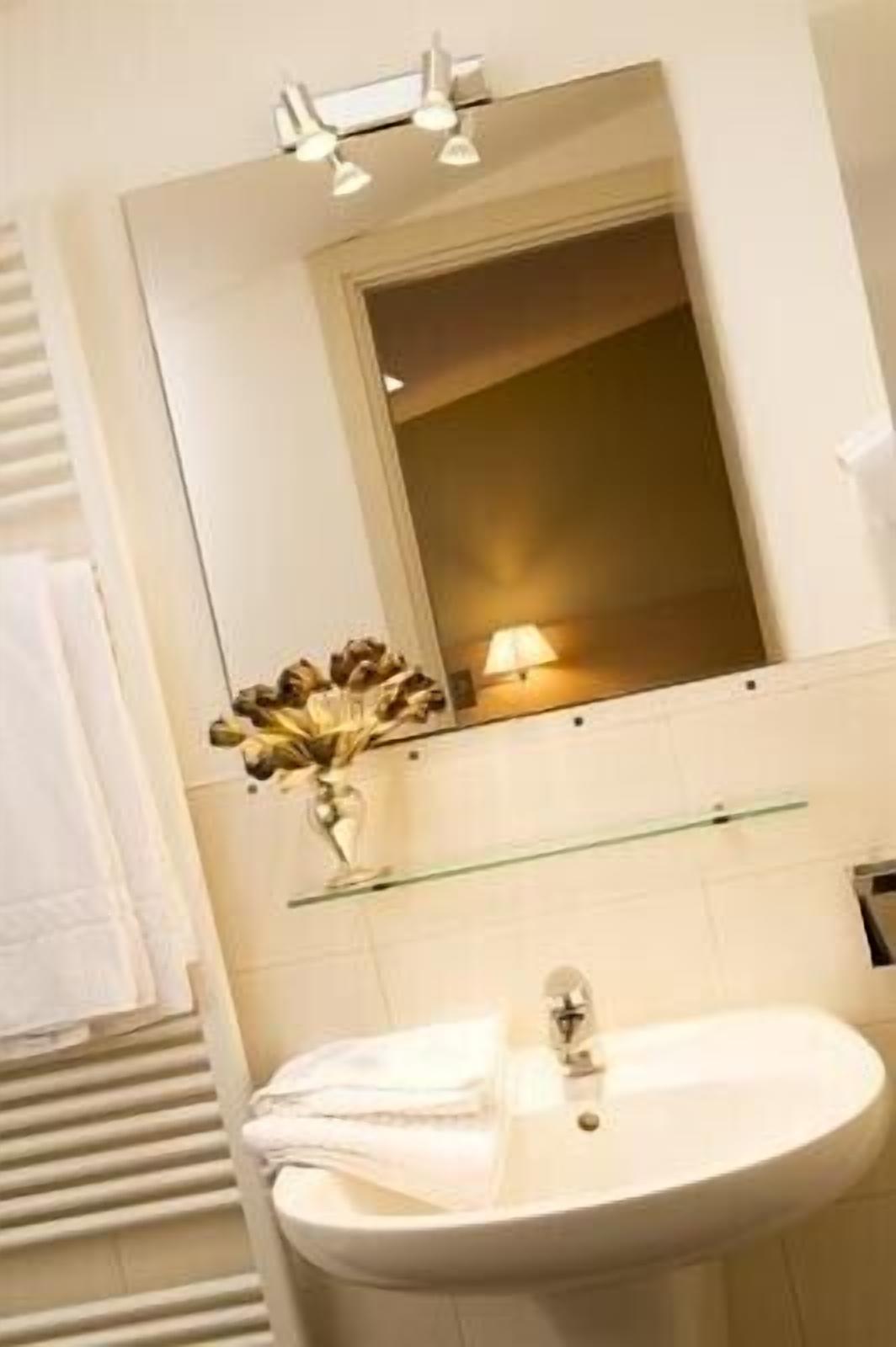 Une salle de bain avec un lavabo blanc, un miroir au-dessus et un vase rempli de fleurs posé sur une étagère.