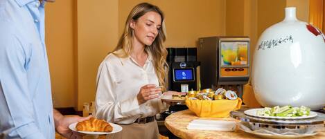 Una escena de desayuno de hotel con una mesa de buffet, una máquina de café y un gran jarrón decorativo.