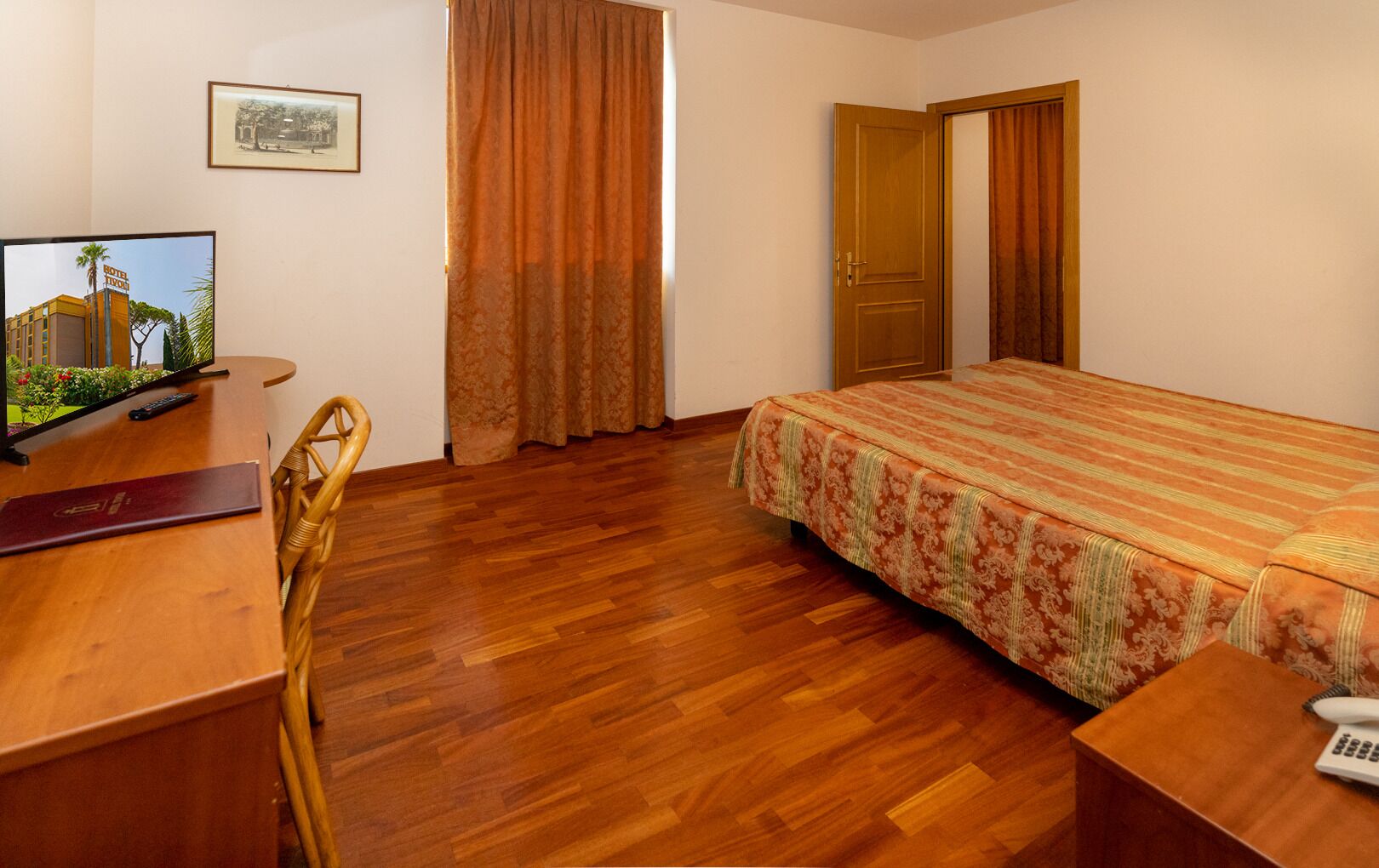 A hotel room with a wooden floor, a bed with a floral bedspread, a desk with a remote control, a chair, and a door.