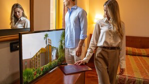 A hotel room with a large flat-screen TV on a wooden desk, a bed with a patterned bedspread, and a nightstand with a lamp. - Hotel Tivoli (Tivoli)