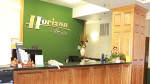 A reception desk with a wooden finish, a green wall with the Horizon Inn & Suites name, a reception counter, a desk lamp, a telephone, and a small plant.