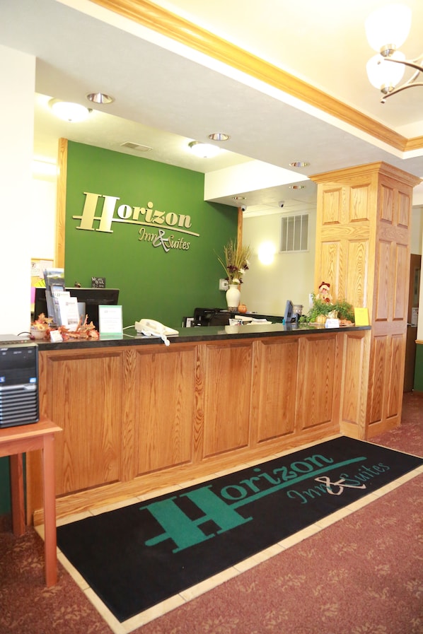 A reception desk with a wooden finish, a green wall with the Horizon Inn & Suites name, a reception counter, a desk lamp, a telephone, and a small plant. - Horizon Inn and Suites (West Point)
