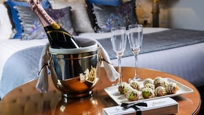 Een bed met een gestoffeerd hoofdbord, een nachtkastje met een champagnefles, twee glazen en een bord met versierde lekkernijen.