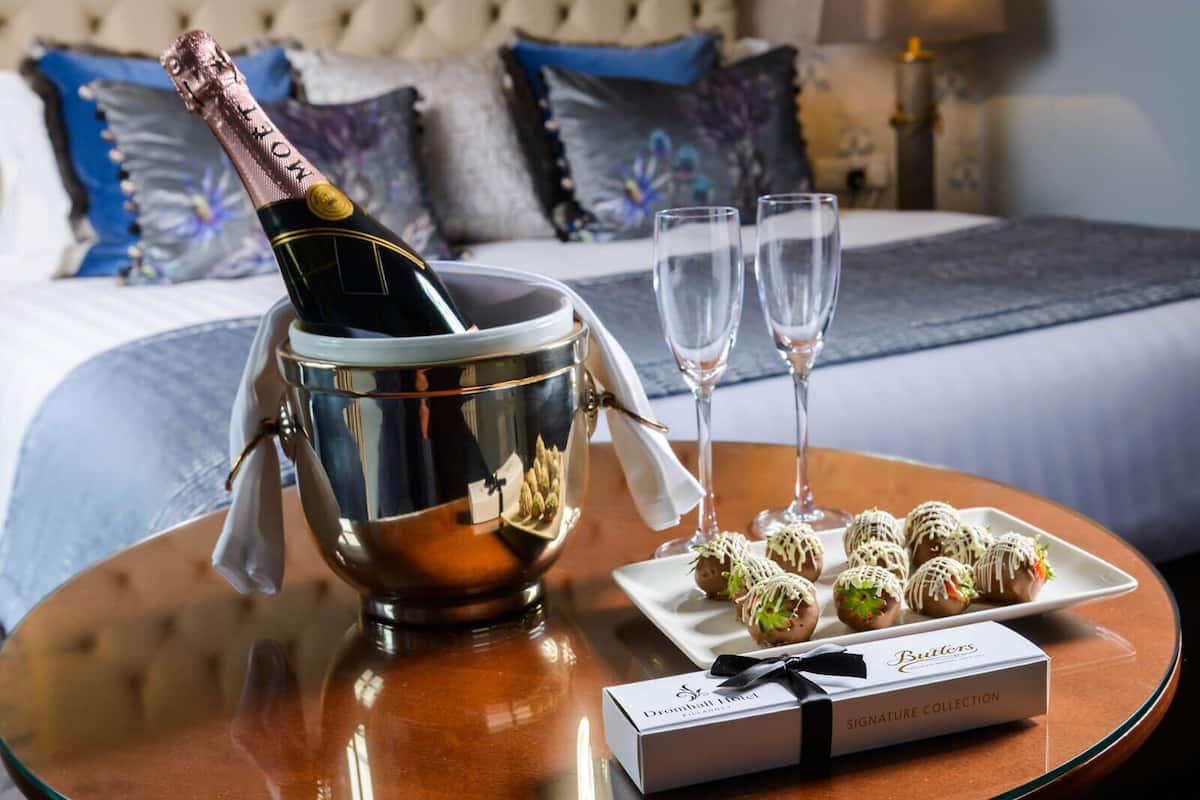 Ein Bett mit einer gepolsterten Kopfteile, ein Nachttisch mit einer Champagnerflasche, zwei Gläsern und einer Platte mit verzierten Köstlichkeiten.