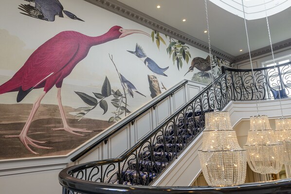 Una imponente escalera con barandales negros decorados y un mural en la pared con pájaros y follaje.
