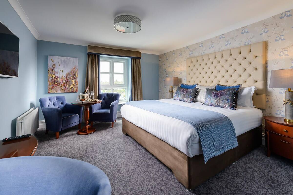 Ein Hotelzimmer mit einem großen Bett, einem gepolsterten Kopfteil, einer Sitzecke mit Sessel und Tisch sowie einem Fenster mit Vorhängen.