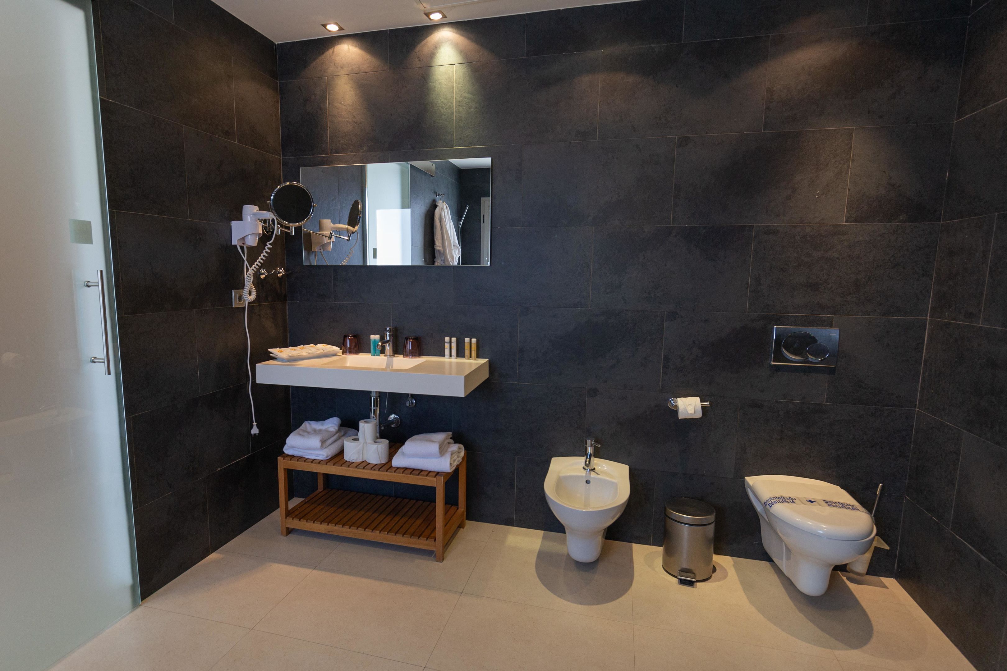 Une salle de bain moderne avec des murs recouverts de carreaux foncés, un meuble-lavabo en bois, un bidet et des toilettes.