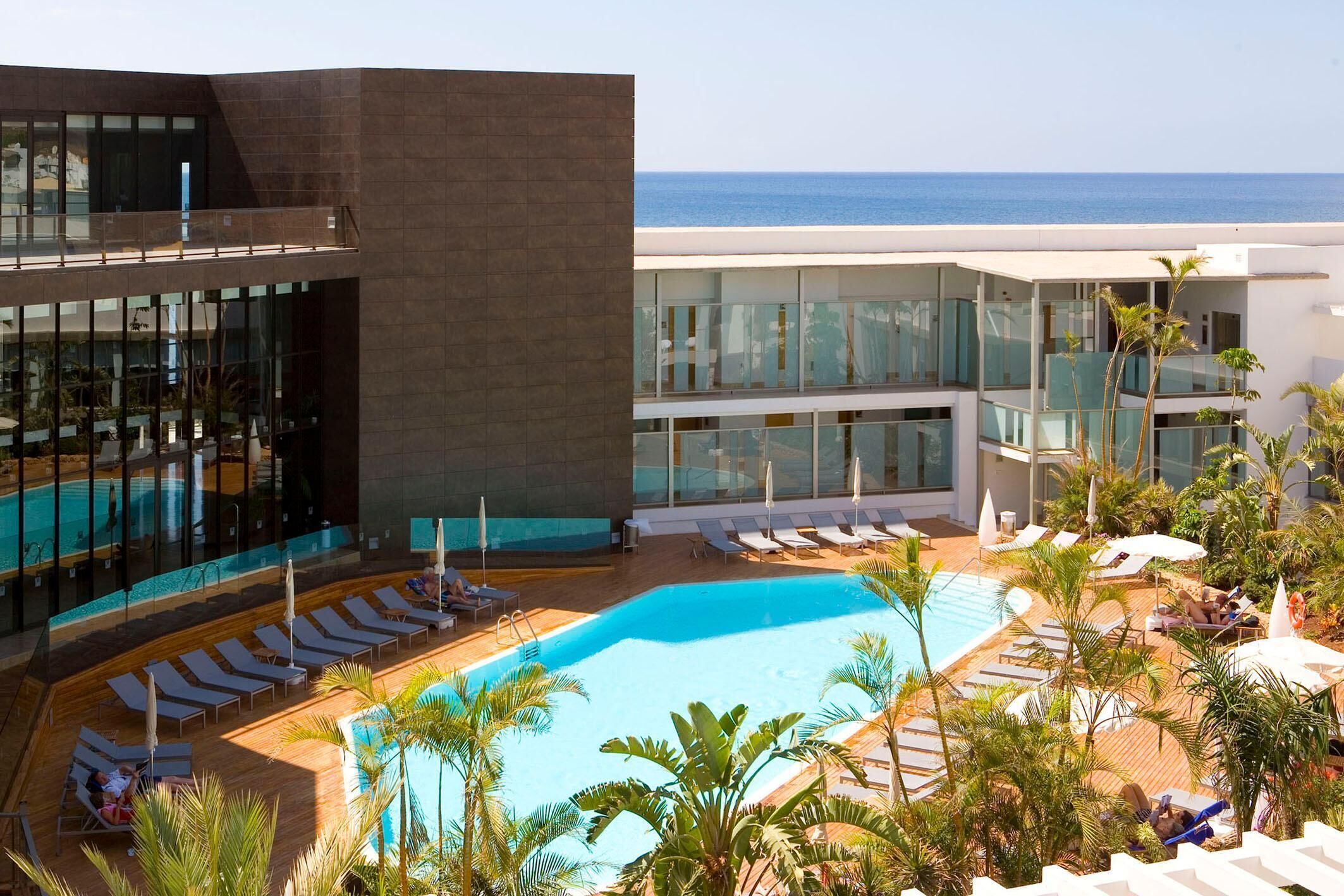 Un hotel moderno con zona de piscina, sillas de descanso y vista al mar.