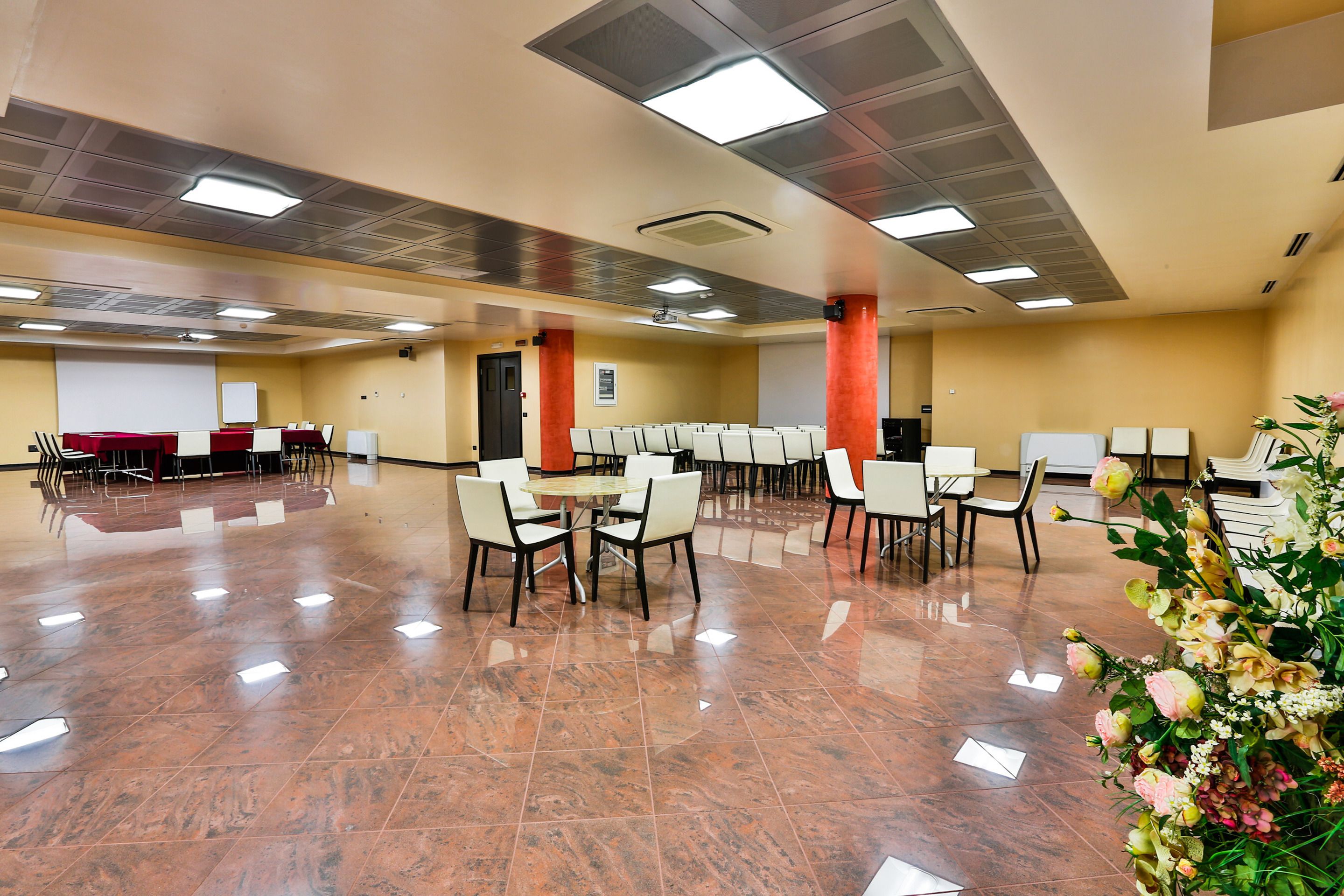 Una amplia sala de conferencias con piso de baldosas pulidas, varias mesas y sillas, y una pantalla grande al frente.