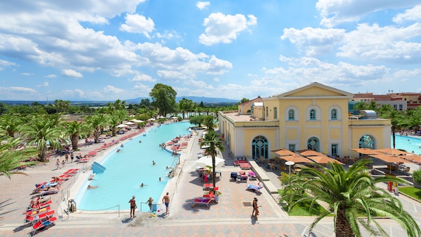 A large swimming pool with lounge chairs and umbrellas, surrounded by palm trees and a building with arched windows. - Victoria Terme Hotel (Tivoli)
