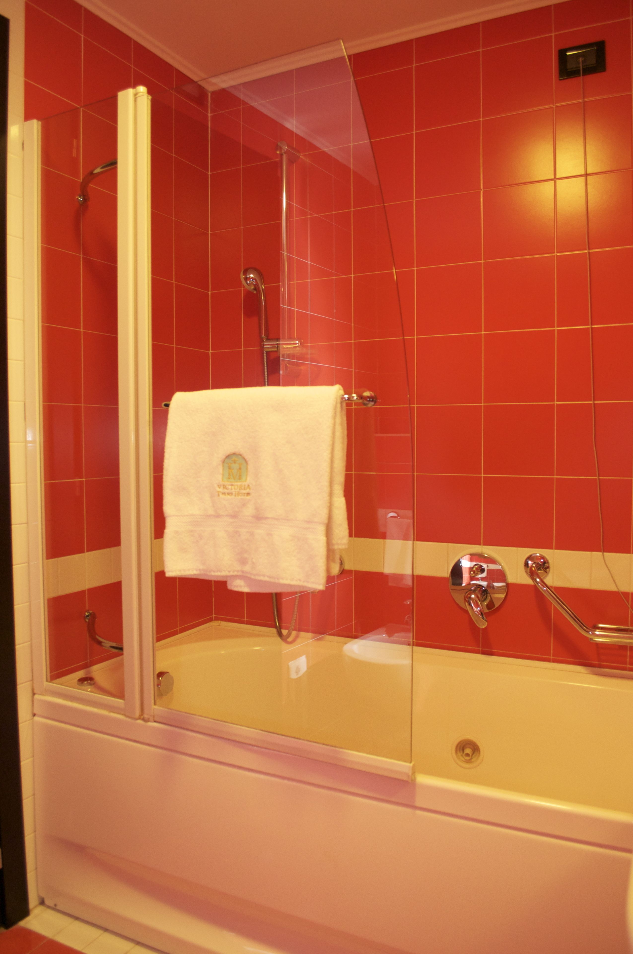 A bathroom with a glass-enclosed shower, a white bathtub, and red tiled walls.