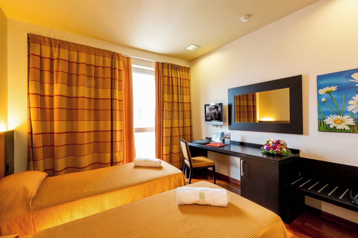 A hotel room with two beds, a desk, a chair, a desk lamp, a mirror, and a painting on the wall.