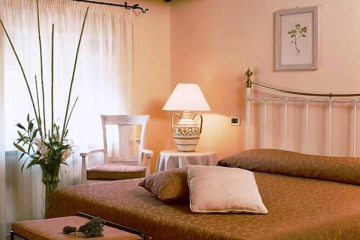 Une chambre d’hôtel avec un lit, une chaise, une table avec une lampe, un vase de fleurs et un radiateur.