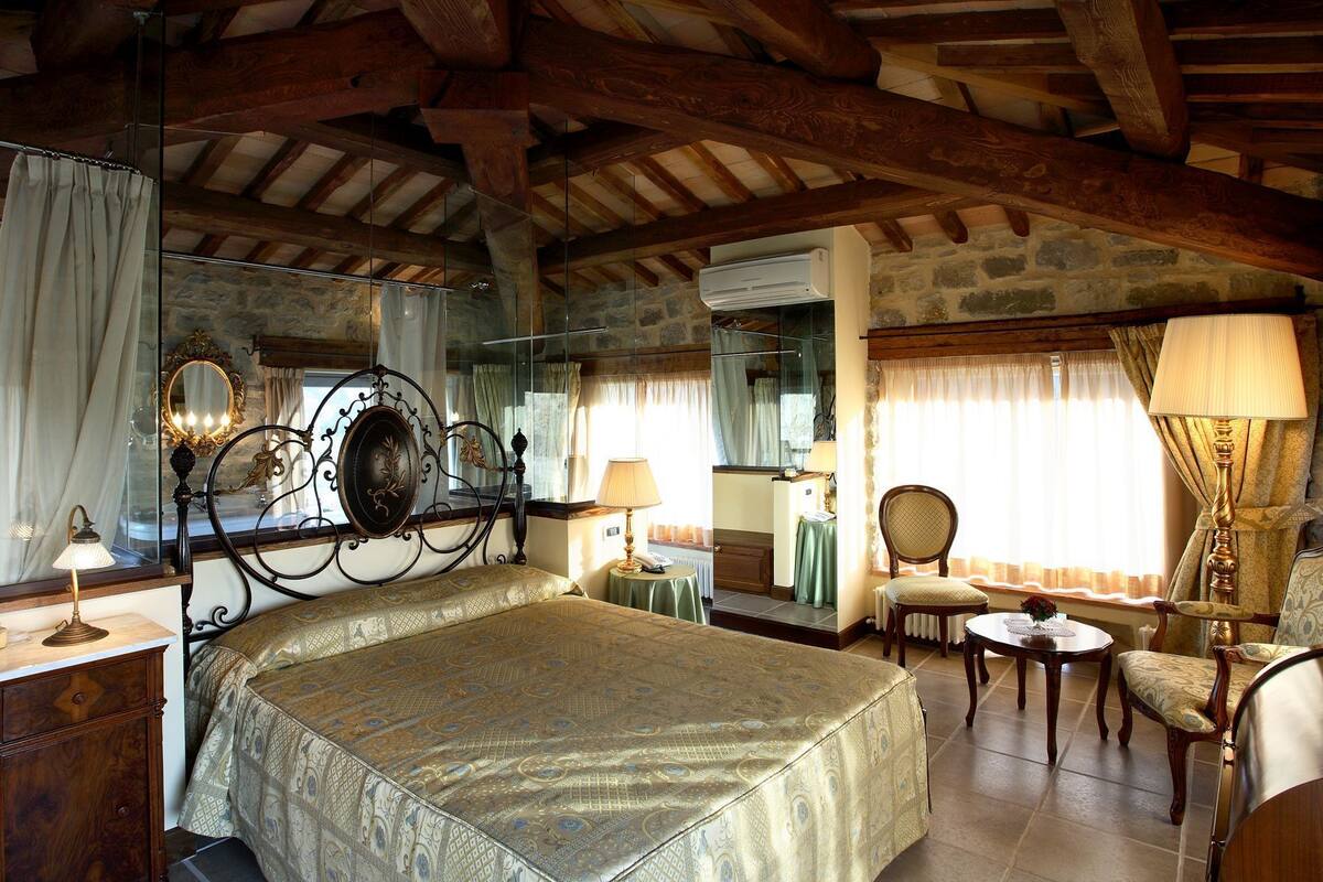 Une chambre à coucher avec un lit, un miroir, une commode, une chaise, une table et une fenêtre avec des rideaux.