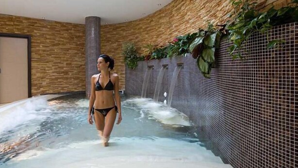 Une femme se promène dans un espace de détente avec un jacuzzi et un mur recouvert de carreaux et orné de plantes.
