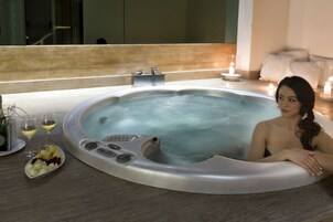Una bañera de hidromasaje con una mujer sentada en su interior, rodeada de velas y una bandeja de frutas.
