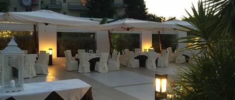 An outdoor dining area with round tables covered in white tablecloths and chairs, under white canopies.