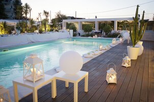 Un área junto a la piscina con cubierta de madera, sillas reclinables blancas y faroles.
