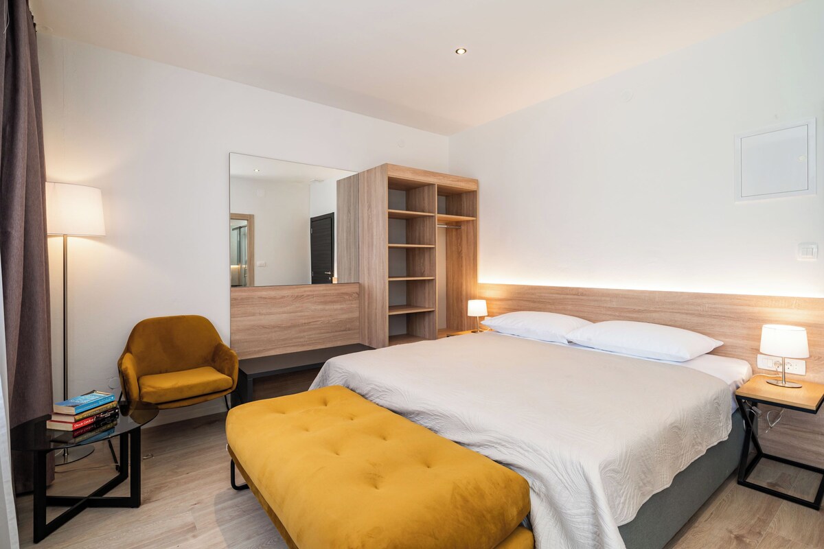 A hotel room with a bed, a bedside table, a chair, a bench, a wardrobe, and a mirror.