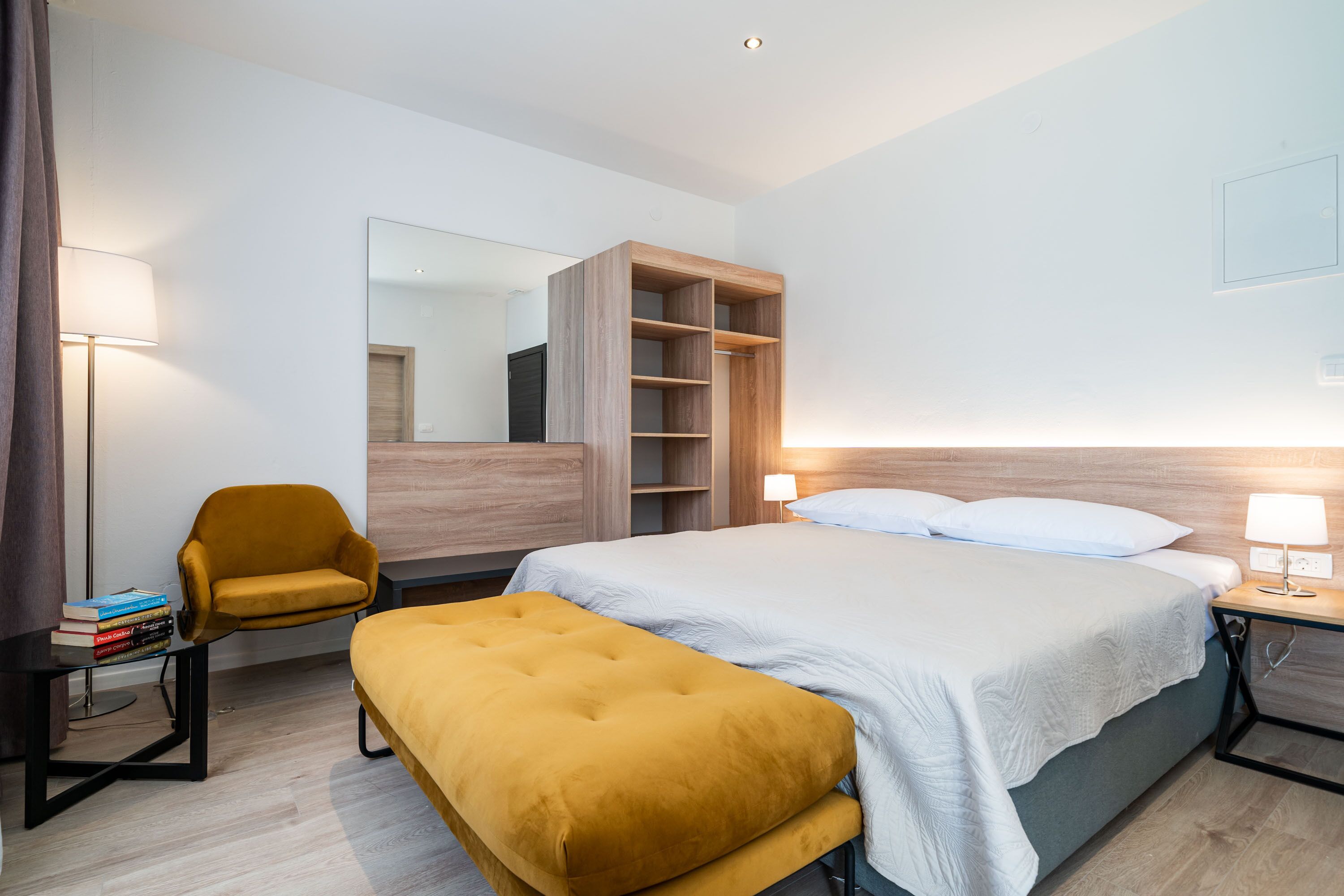 A modern hotel room with a bed, a bedside table with a lamp, a wooden wardrobe, a yellow armchair, and a small table with books.