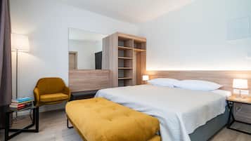 A modern hotel room with a bed, a bedside table with a lamp, a wooden wardrobe, a yellow armchair, and a small table with books.