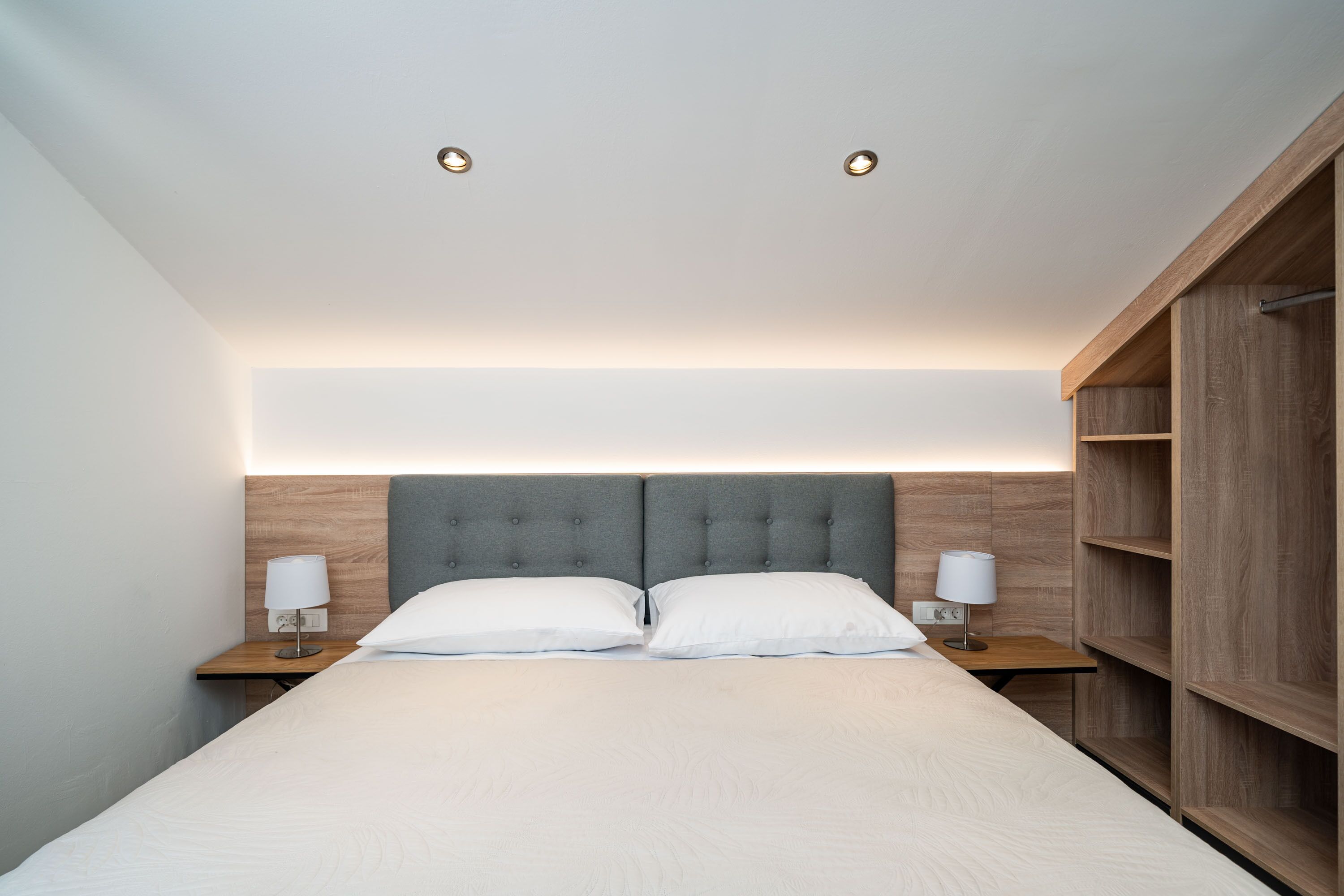 A modern bedroom with a large bed, two bedside tables with lamps, a wooden headboard, and built-in shelving.