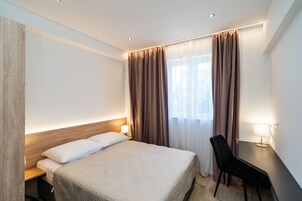 Habitación de hotel con una cama grande, un escritorio con una silla, una lámpara y una ventana con cortinas.