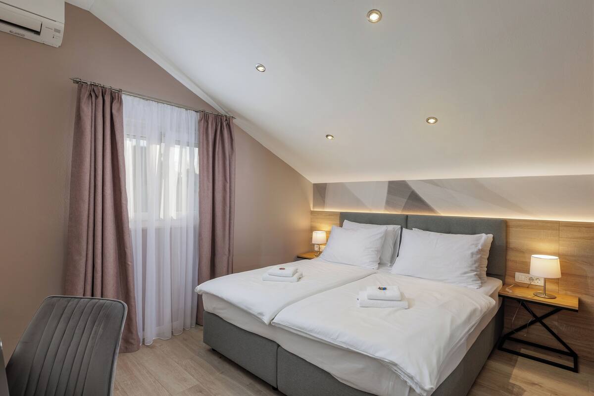 Habitación de hotel con una cama, dos lámparas de noche, una silla y una ventana con cortinas.