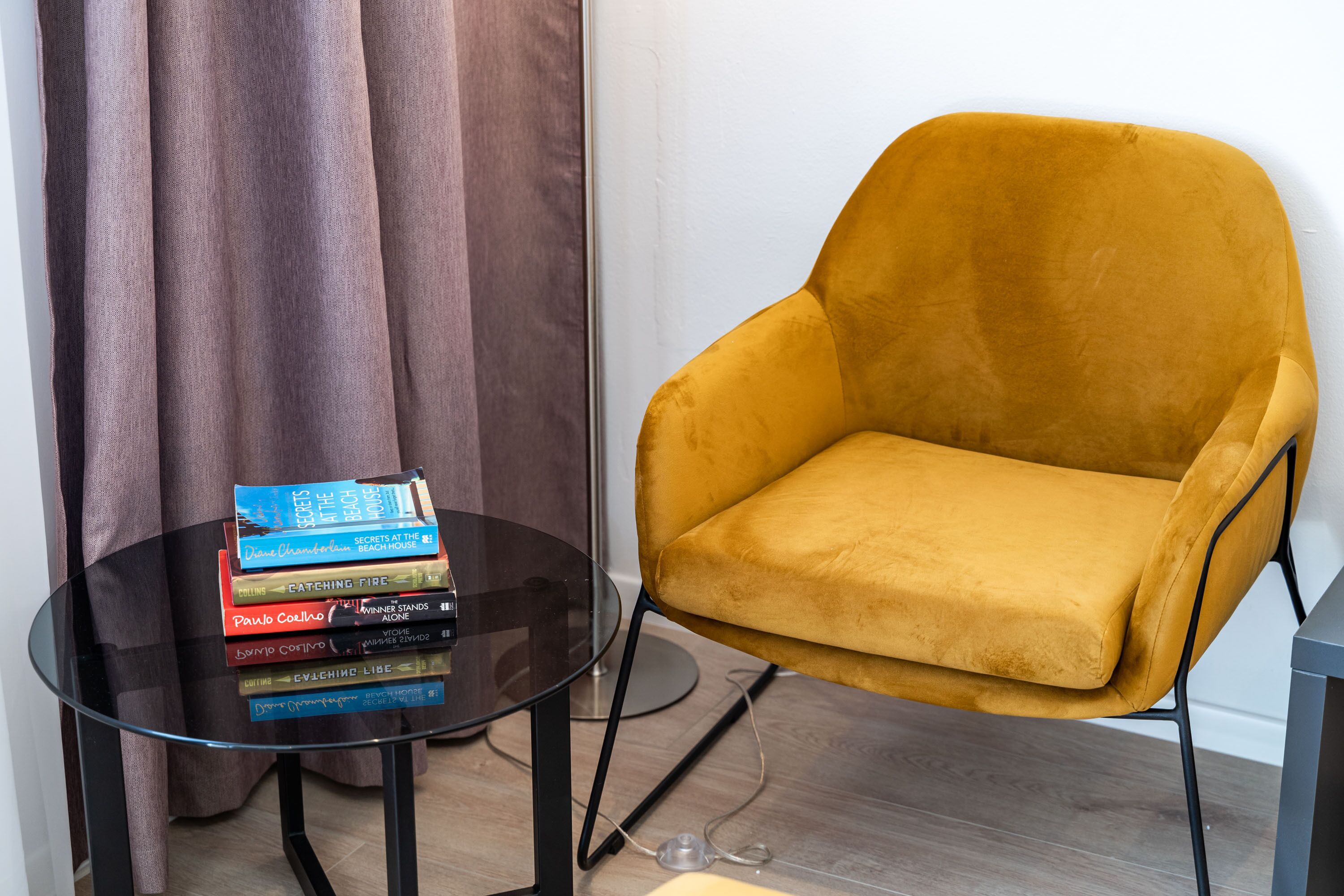 A room with a yellow armchair, a glass table with books, and a curtain.