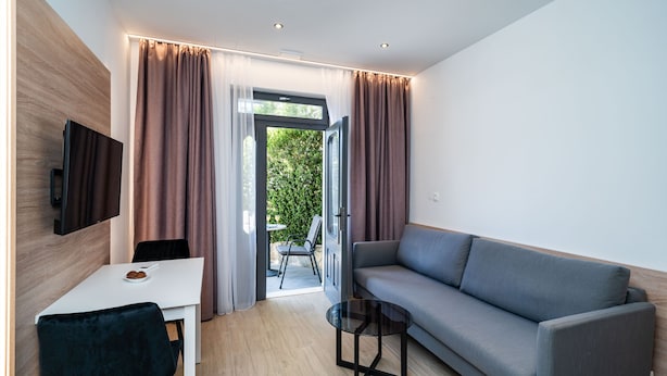 Una sala de estar moderna con un sofá gris, una mesa blanca y una silla negra. Hay un televisor montado en la pared y una puerta de cristal que da al exterior.