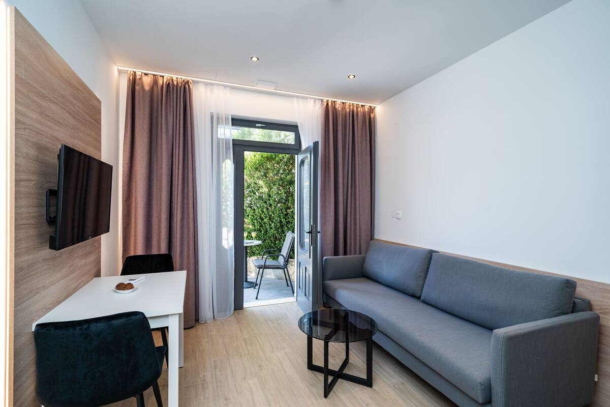 Una sala de estar moderna con un sofá gris, una mesa blanca y una silla negra. Hay un televisor montado en la pared y una puerta de cristal que da al exterior.