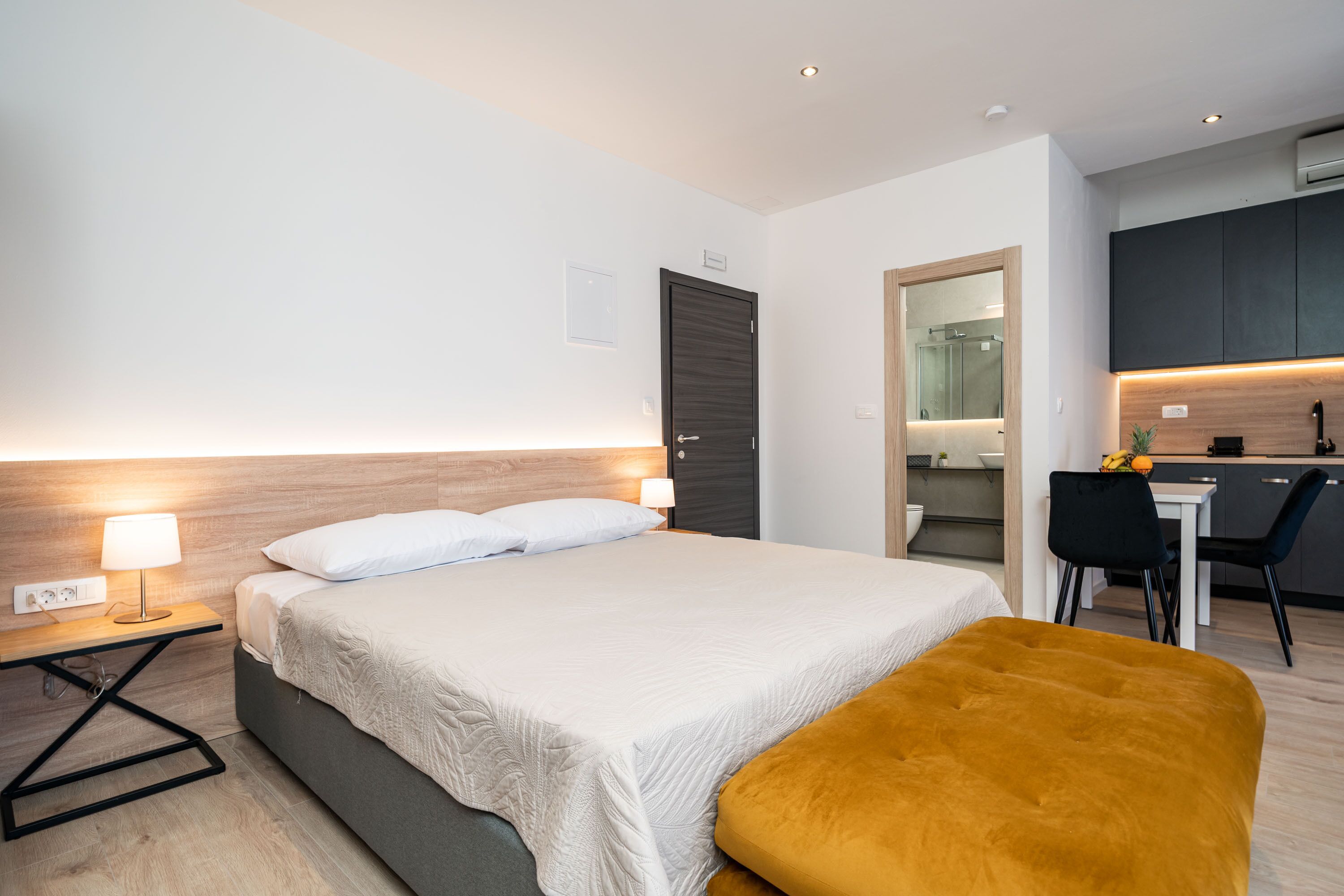 A modern hotel room with a bed, bedside table, lamp, and a small desk area with a chair.