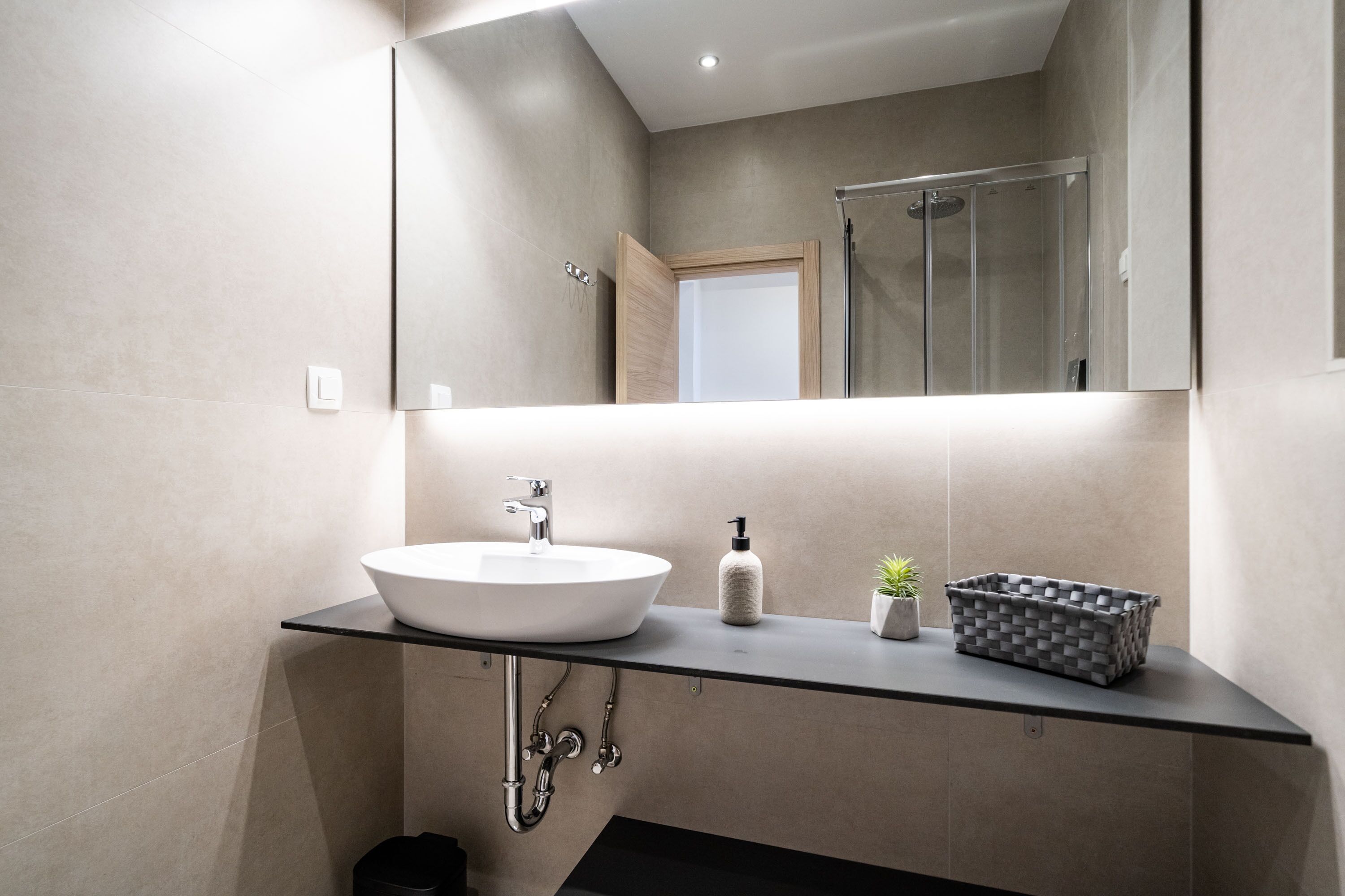 A modern bathroom with a large mirror, a round sink, a shower enclosure, and a small potted plant.